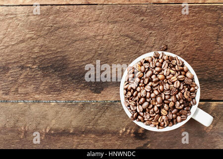 Overhead Schuss einer weißen Tasse gefüllt mit ganz frischen Kaffeebohnen über eine hölzerne Tischplatte. Flache Laien Draufsicht Stil. Stockfoto