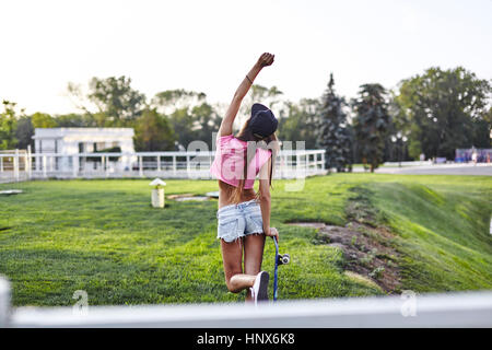 Junge Frau, die zu Fuß durch den Park, die Skateboard, Stanzen Luft, Rückansicht Stockfoto