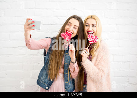 Stilvolle Freundinnen machen Selfie Foto mit roten Bonbons auf der weißen Wand-Hintergrund Stockfoto