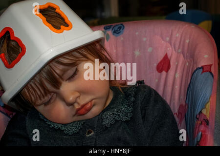 Blonde Mädchen schlafend auf einer Babyschaukel mit einem Kunststoff-Spielzeug-Box auf dem Kopf als Hut. Sie trägt eine schwarze Spitzenbluse Stockfoto