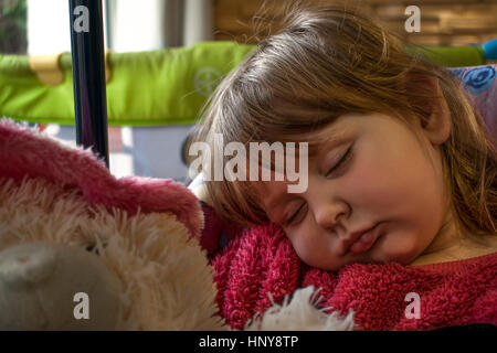 Blondes Babymädchen trägt einen rosa Fell Pullover schlafen auf einem Stuhl mit einem pelzigen Teddy-Bären auf dem Schoß. Stockfoto