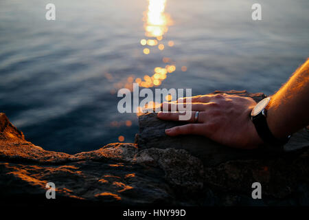 Schuss von Herren linke Hand auf einem Felsen, trägt eine einfache Ehering und eine Uhr hautnah. Meer und Lichtspuren auf dem Hintergrund. Stockfoto