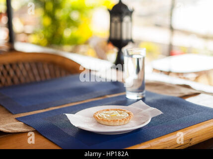 Zitronentorte mit Creme-Füllung und Zitronenscheiben auf einem weißen Teller, blauen Matte und Holz Café-Tisch Stockfoto