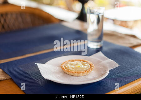 Zitronentorte mit Creme-Füllung und Zitronenscheiben auf einem weißen Teller, blauen Matte und Holz Café-Tisch Stockfoto