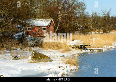 Roten hölzernen Bootsschuppen in Winterlandschaft. Hölzerne Pier und Schilf am Ufer. Wald im Hintergrund. Boot an Land unter Schnee. Lage Sjodala in Bleki Stockfoto