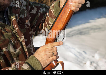 Mann angeklagt Windjacke im tief verschneiten Winterwald.  Ein Mann in eine Camouflage-Jacke hält ein Luftgewehr und eine Schachtel mit Schrot, Munition. Stockfoto