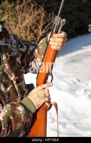 Mann angeklagt Windjacke im tief verschneiten Winterwald.  Ein Mann in eine Camouflage-Jacke hält ein Luftgewehr und eine Schachtel mit Schrot, Munition. Stockfoto