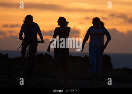 Drei Freundinnen auf einer Küstenstraße bei Sonnenuntergang, spazieren und Rad fahren, Silhouette am Abendhimmel. Osterinsel Stockfoto