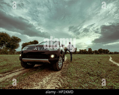 Ein Mitsubishi Pajero Sport SUV-Fahrzeug in ein grünes Feld mit Wanderwegen, Lichter auf, dramatische Wolken am Himmel, und siehe, ein Mann stand ihm zurück Übersicht Stockfoto