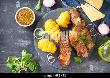 BBQ Schweinerippchen mit Senf. Fried in Scheiben geschnitten Grill Schweinerippchen und gebackene Kartoffel mit Käse mit einer Vielzahl von Gemüse und grünen auf Schiefer Hintergrund Stockfoto