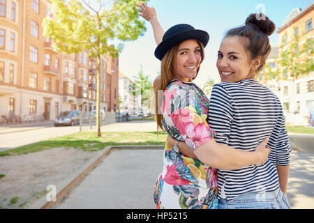 Glücklich freundlich trendige junge Frauen gehen Arm in Arm entlang einer ruhigen städtischen Straße wellenartig bewegen und drehen Blick zurück auf die Kamera, mit Textfreiraum Stockfoto