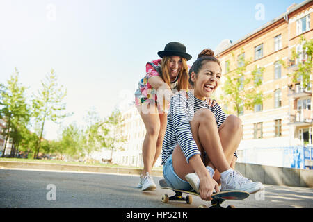 Niedrigen Winkel Blick auf glückliche teenaged Frau lachend Freund auf Skateboard unterwegs im Stadtgebiet mit Apartments, die um sie herum schieben Stockfoto