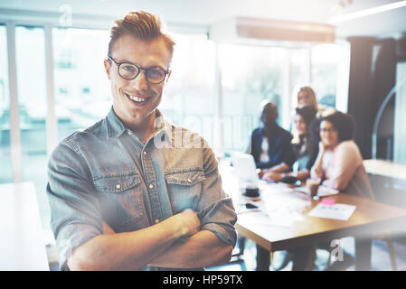 Selbst sicher Jungunternehmer in Gläsern und Freizeitkleidung stehend Arme gefaltet und lächelnd in hellen Büro, Blick in die Kamera. Sein Team ist Unschärfe Stockfoto