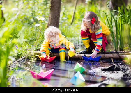 Kinder spielen mit bunten Papierschiffchen in einem kleinen Fluss an einem sonnigen Frühlingstag. Kinder spielen, die Natur zu erforschen. Bruder und Schwester, die Spaß an einem Stockfoto