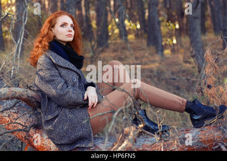 Schöne elegante rothaarige Frau sitzend auf Baumstamm im herbstlichen Wald. Rothaarige Mädchen im Herbst Kleidung in den Wald. Rothaarige Frau outdoor im Herbst park Stockfoto