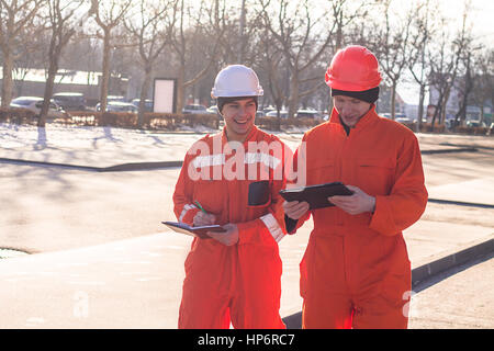 Team von jungen Ingenieuren Projekt diskutieren und schreiben in das Notizbuch. Der Verschleiß Overalls und Helme. Straße Hintergrund Stockfoto