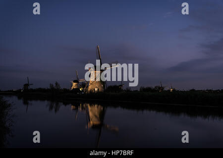 Mehrere beleuchtete Windmühlen mit Spiegelbild im Wasser des Flusses in der Dämmerung am Anfang der Nacht in Kinderdijk, Niederlande. Stockfoto