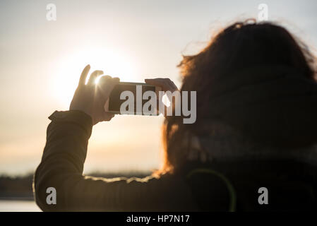 junge Frau, die Sonne mit dem Handy zu fotografieren. Menschen, die gefrorenen Strand im Winter kalt genießen. Wandern und entspannen Stockfoto