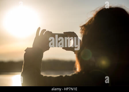 junge Frau, die Sonne mit dem Handy zu fotografieren. Menschen, die gefrorenen Strand im Winter kalt genießen. Wandern und entspannen Stockfoto