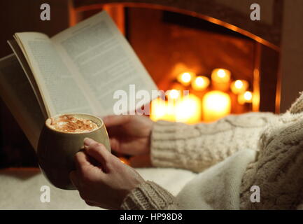 Eine Frau liest ein Buch mit einem Becher Sahne gekrönt Mokka (Kaffee und Schokolade) am offenen Kamin beleuchtet mit Kerzen in einem gemütlichen englischen Haus im winter Stockfoto