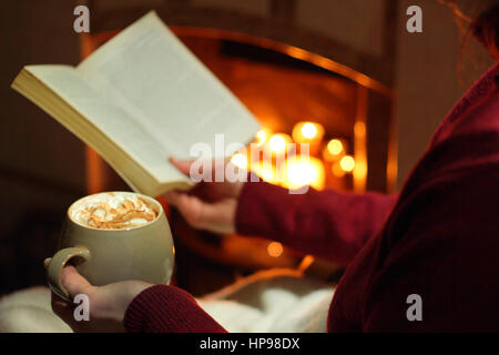 Eine Frau liest ein Buch mit einem Becher Sahne gekrönt Mokka (Kaffee und Schokolade) am offenen Kamin beleuchtet mit Kerzen in einem gemütlichen englischen Haus im winter Stockfoto