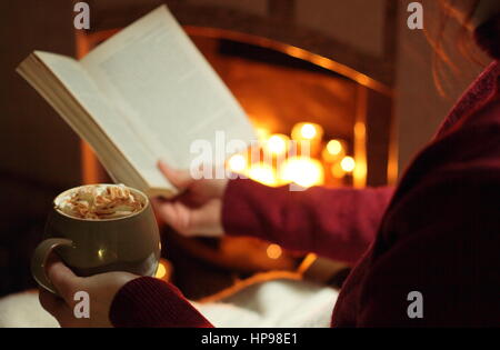 Eine Frau liest, dass ein Buch mit einem Becher Sahne gekrönt Mokka (Kaffee und Schokolade) am offenen Kamin beleuchtet mit Kerzen in einem cos englischen Haus im winter Stockfoto