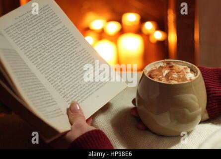 Eine Frau liest ein Buch mit einem Becher Sahne gekrönt Mokka (Kaffee und Schokolade) am offenen Kamin beleuchtet mit Kerzen in einem gemütlichen englischen Haus im winter Stockfoto