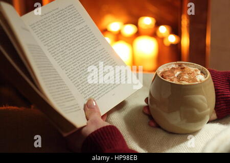 Eine Frau liest ein Buch mit einem Becher Sahne gekrönt Mokka (Kaffee und Schokolade) am offenen Kamin beleuchtet mit Kerzen in einem gemütlichen englischen Haus im winter Stockfoto