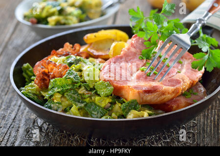 Geräucherter Nacken vom Schwein mit Wirsing, Speck und Bratkartoffeln serviert in einer Eisenpfanne auf einem Holztisch Stockfoto