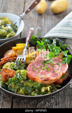 Geräucherter Nacken vom Schwein mit Wirsing, Speck und Bratkartoffeln serviert in einer Eisenpfanne auf einem Holztisch Stockfoto