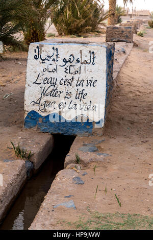 Merzouga, Marokko.  Bewässerungskanal Wassertragen Landwirte Plots in der Oase Merzouga.  "Wasser ist Leben" in fünf Sprachen. Stockfoto