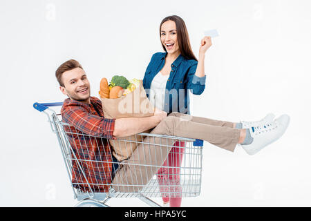 man sitzt im Einkaufswagen mit Einkaufstüte und lächelnde Frau leere Karte zeigen Stockfoto