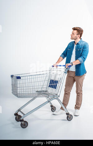 hübscher junger Mann stehend mit Einkaufswagen und wegsehen Stockfoto