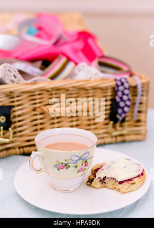 Cream Tea. Tassen Tee und Scones mit Marmelade und Clotted Cream, begleitet von einem Weidenkorb gefüllt mit Stoff Stockfoto