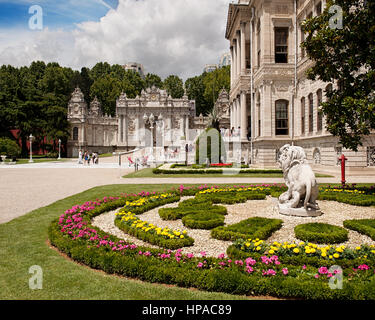 Reich verzierte Gärten des Dolmabahce Palace mit gepflegten Blumenbeeten, einer Löwenstatue und dem großen Kaisertor unter einem teilweise bewölkten Himmel. Stockfoto