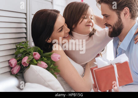 Feierlichkeiten zum Tag der Mütter. Freuen uns angenehme positive Mädchen umarmt ihre Eltern und ihr Vater zu betrachten, und dabei Spaß haben Stockfoto