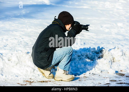 Jungen Erwachsenen Fotografen kniend im Winter Umgebung zu fotografieren. Stockfoto