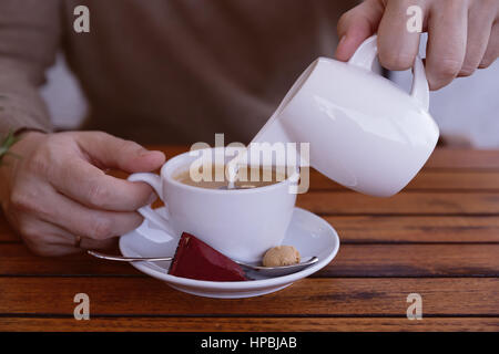 Geschäftsmann in Pulover Gießen Milch oder Sahne Tasse Kaffee im Café-Bar oder im Restaurant auf dem Holztisch. Close-up. Man gießt Sahne in weiße Tasse Stockfoto