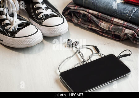 Kleidung & Zubehör: Hipster Turnschuhe, Jeans, kariertes Hemd, Cowboy Gürtel, Telefon mit Kopfhörern auf hölzernen Hintergrund. Reisen-Konzept Stockfoto