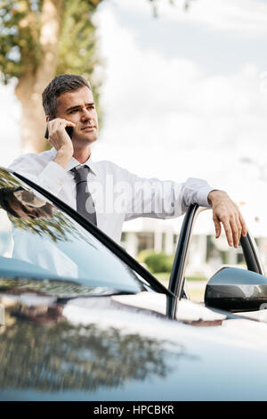 Zuversichtlich Geschäftsmann mit einem Telefonat mit seinem Smartphone und stützte sich auf die Autotür Stockfoto