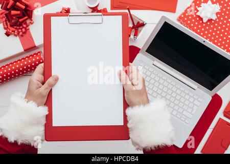 Planung für Weihnachten Weihnachtsmann schreibt auf einem Klemmbrett, er mit einem Laptop und Verpackung Geschenke, leere Kopie Raum, Ansicht von oben Stockfoto