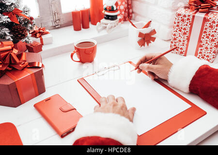 Santa Claus am Schreibtisch arbeiten und schreiben auf ein leeres Blatt auf einem Klemmbrett, ist er immer bereit für Weihnachten Stockfoto