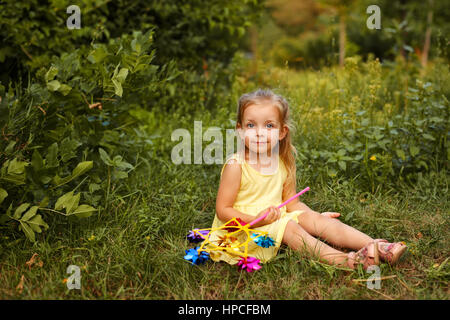 Niedliche kleine Mädchen hält ein Windrad. Sie sitzt auf dem Rasen. Ein Spaziergang im Sommerpark. Sommer-Urlaub. Outdoor-Aktivitäten. Stockfoto