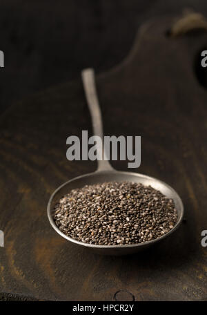 Rohe unbearbeitete schwarz Chia-Samen in Metalllöffel auf Holzbrett auf dunklem Hintergrund Stockfoto