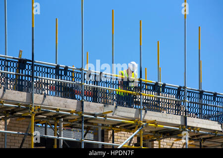 Bauarbeiter auf Gerüsten auf neue Baustelle legen Ziegelsteine hinter Sicherheit netting, umrahmt von blauen Sommerhimmel Stockfoto