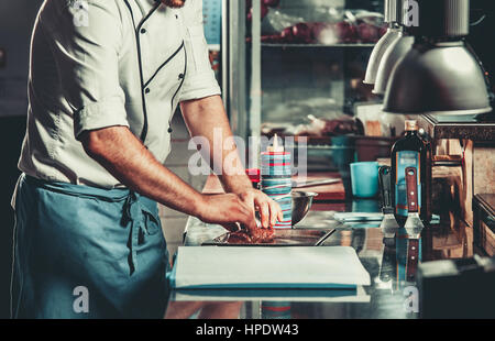 Junge weiße Koch in der Küche Interieur. Mann marinieren Rindersteak auf einem Tablett. Fleisch für den Grill bereit und dienen. Nur Hände Stockfoto