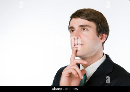 Kopf und Schulter Blick auf dunklen Anzug eines jungen Mannes vor weißem Hintergrund den Zeigefinger auf das Kinn platziert mit nachdenklichen Blick nach links oben. Stockfoto