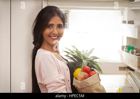 Lächelnde junge Frau hält Obst Tasche stehen vor offenen Kühlschrank Stockfoto