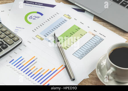 Moderner Arbeitsplatz im Büro: Ausdrucke von Diagrammen und Finanzdaten neben einem Laptop, Kaffee und Rechner auf dem Schreibtisch (reduzierte Ton) Stockfoto