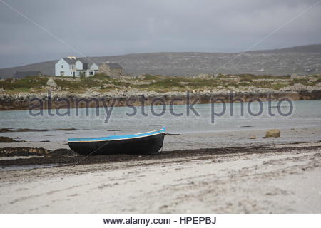 Ein blau lackierten Currach Boot liegt gefesselt am Ufer in Connemara, Co. Galway, Irland mit einem Haus im Hintergrund zu sehen Stockfoto